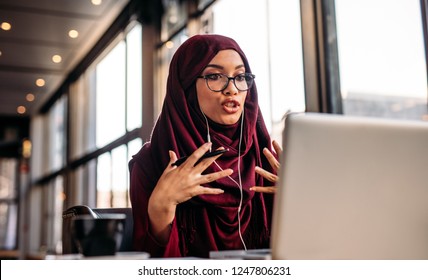Businesswoman in hijab having a video chat on laptop while sitting at coffee shop. Female sitting at cafe and making video call using earphones and laptop computer.