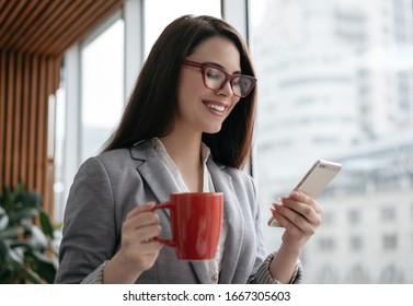 Businesswoman Holding Cellphone And Cup Of Coffee, Communication, Checking Email In Office. Beautiful Woman In Stylish Red Eyeglasses Using Mobile App For Online Shopping. Freelancer Receive Payment