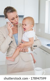 Businesswoman Holding Her Crying Baby Talking On The Phone At Home In The Kitchen