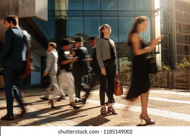 Businesswoman holding her hand bag standing still on a busy street with people walking past her using mobile phones. Woman standing amidst a busy office going crowd hooked to their mobile phones.