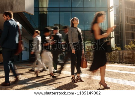 Businesswoman holding her hand bag standing still on a busy street with people walking past her using mobile phones. Woman standing amidst a busy office going crowd hooked to their mobile phones.
