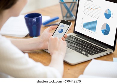 Businesswoman holding smartphone while working with laptop at home office desk, using cross platform responsive design apps for project analysis, analyzing cloud data statistics, close up rear view - Powered by Shutterstock