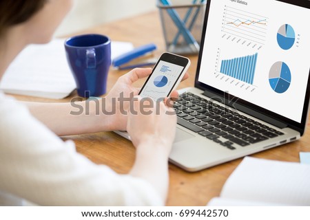 Businesswoman holding smartphone while working with laptop at home office desk, using cross platform responsive design apps for project analysis, analyzing cloud data statistics, close up rear view