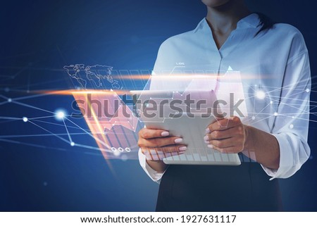 Businesswoman holding tablet and using technological approach to optimize business process. System hologram charts and graph flying nearby smartphone. Office on background.
