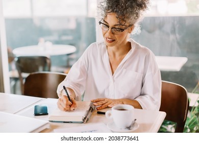 Businesswoman making notes in her work journal while working in a cafe. Mature businesswoman smiling cheerfully while sitting alone at a cafe table. Businesswoman planing her week.