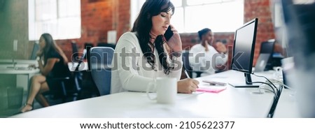 Businesswoman making notes while speaking on a phone call. Creative businesswoman connecting with her business clients in a modern office. Young businesswoman making business plans.