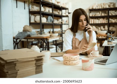 Businesswoman making a phone call while using laptop in ceramic workshop - Powered by Shutterstock