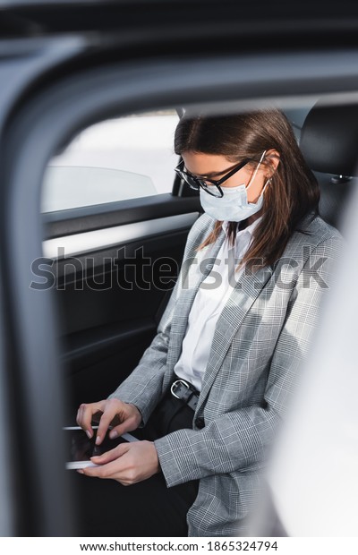 businesswoman in medical mask using digital\
tablet while riding in car on blurred\
foreground