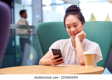 Businesswoman With Mobile Phone Working At Table In Breakout Area Of Office Building