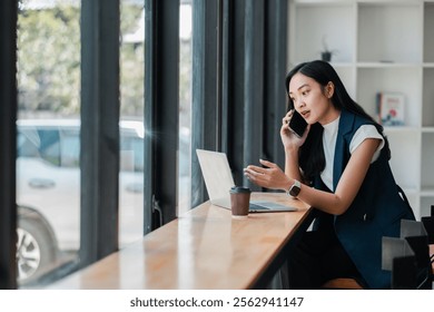 Businesswoman multitasking in a cafe, using a laptop and phone. Bright, modern setting with natural light. - Powered by Shutterstock