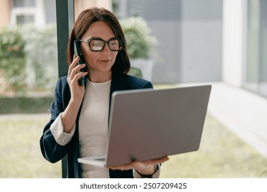 A businesswoman multitasking talking on phone, working on laptop in modern office - Powered by Shutterstock