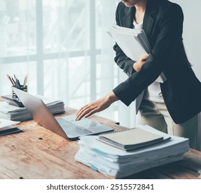 Businesswoman Navigating a Mountain of Work: A professional woman expertly navigates a cluttered desk, her focused gaze directed at a laptop amidst a towering stack of paperwork, showcasing the demand - Powered by Shutterstock