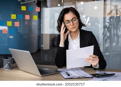 Businesswoman in office analyzing documents, discussing on smartphone, multitasking with laptop, focusing on reports. Concept of business analysis, stress, office work, professional life - Powered by Shutterstock