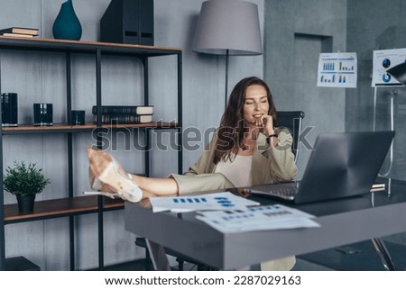 Businesswoman in office at her desk with her feets up on her desk and looking at screen with smile