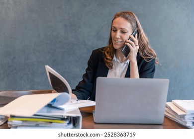 Businesswoman on Phone Call Reviewing Documents: Multitasking, Communication, and Office Work with Laptop and Papers - Powered by Shutterstock