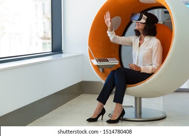 Businesswoman playing virtual game in studio. Woman in office clothes and virtual reality glasses sitting in interactive chair with laptop and touching air. VR game concept