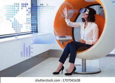 Businesswoman playing VR game and virtual statistic graphics. Woman in office clothes and virtual reality glasses sitting in interactive chair with laptop and touching air. VR game concept - Powered by Shutterstock