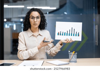 Businesswoman presenting data analysis during video call, holding bar chart with confused expression while explaining statistics. Concept of business presentation, communication, analysis. - Powered by Shutterstock