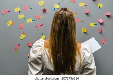 Businesswoman Ready For Her Next Task, She's Looking At The Impressive Wall Of Assignments, Her Back Is Turned, She's Holding Papers In One Arm, Pen In The Other.