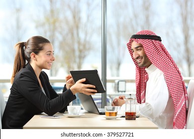 Businesswoman or saleswoman working with an arab man showing products in a tablet in a coffee shop - Powered by Shutterstock