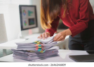 Businesswoman searching through large stack of paperwork with colorful sticky notes attached at her office desk with computer screen showing graphs in background