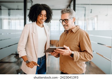 A businesswoman and a senior male colleague collaboratively review content on a tablet in a bright office space, reflecting a multigenerational workplace where experience meets new technology - Powered by Shutterstock