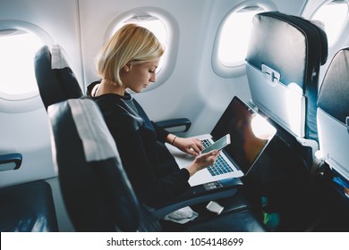 Businesswoman with short haircut sitting in airplane cabin and chatting online on smartphone while checking email on laptop computer with mock up area.Female traveler reading notification on cellular