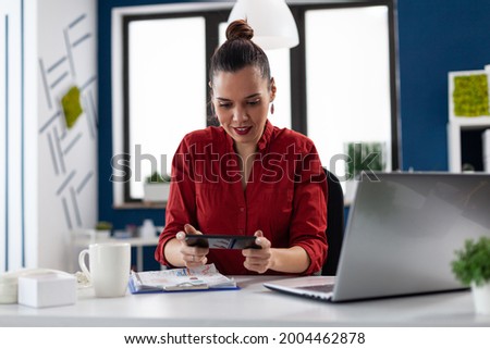 Businesswoman sitting at desk in corporate office playing video games, using smartphone. Entrepeneur taking a break from wwork having fun with games holding phone. Employer having fun at wwork.