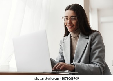Businesswoman sitting in grey coat and glasses and working on computer. Young woman using laptop and smiling, freelancing at co-working space.