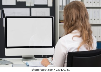 Businesswoman Sitting With Her Back To The Camera Working At A Desktop Computer With The Blank Screen Visible To The Viewer