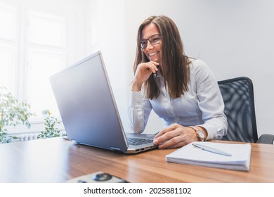 Businesswoman is sitting at her desk, working on her laptop. A woman with glasses is laughing. Sunny office - Shutterstock ID 2025881102