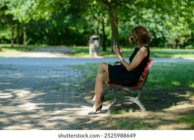 Businesswoman is sitting on a bench, enjoying the summer weather, and using her smartphone. She is wearing a protective face mask - Powered by Shutterstock