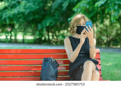 Businesswoman is sitting on a bench in the park, wearing a black face mask and using her smartphone. The woman is working remotely during the coronavirus pandemic - Powered by Shutterstock