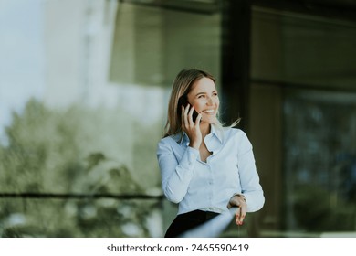 Businesswoman smiles while talking on her smartphone outside a modern office building on a sunny day, dressed in a white blouse and dark skirt - Powered by Shutterstock