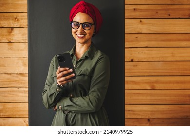 Businesswoman Smiling At The Camera While Holding A Smartphone. Cheerful Muslim Businesswoman Reading A Text Message In An Office. Happy Female Entrepreneur Wearing A Headscarf In A Modern Workplace.