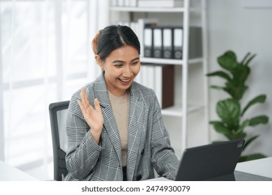 Businesswoman smiling and waving during a video call, working from home office with laptop and bookshelf background.
