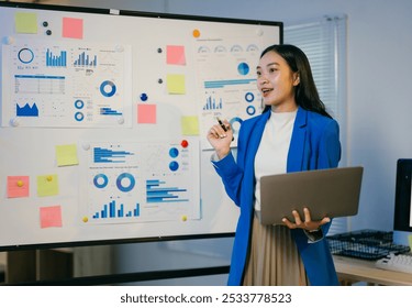 Businesswoman is standing in front of a whiteboard with graphs and charts on it, holding a laptop and giving a presentation