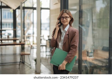 Businesswoman standing and holding a folder while talking smartphone in an office. - Shutterstock ID 2341479237