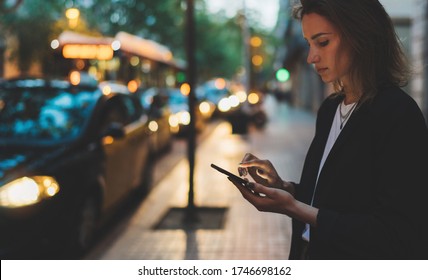 Businesswoman stands by busy road of evening city and calls smartphone of internet technologies, elegant girl waiting for taxi booking hotel on background of passing lights of transport headlights - Powered by Shutterstock