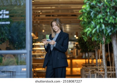 Businesswoman stands in cafe doorway using smartphone for chatting, surfing internet and managing business contracts. Woman cafe owner handles business correspondence and multitasks as manager. - Powered by Shutterstock