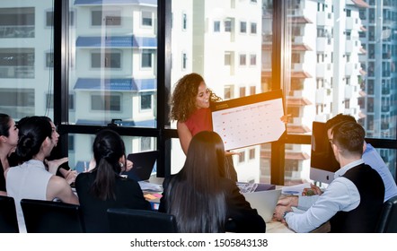 Businesswoman stands to meeting around board table have presenting to colleagues with planning and writing work or vacation on calendar planner.Writing plan on memo scheduled to vacation weekend.Share - Shutterstock ID 1505843138