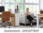 Businesswoman with tablet computer sitting on sofa near water cooler in office