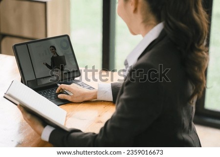 Businesswoman taking notes in a notebook while watching a webinar video course. Serious female student listening to the lecture to study online through e-learning