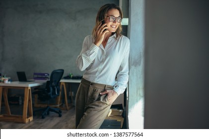 Businesswoman talking on cell phone and smiling. Female entrepreneur standing by a window and talking over mobile phone in office