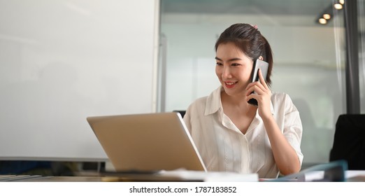 A businesswoman is talking on the mobile phone while sitting and using a computer laptop at the working desk. - Shutterstock ID 1787183852