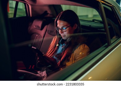 Businesswoman traveling with car on a business trip during night while sitting in a backseat and using a smartphone. Female using mobile phone to send email or messages. View trough car window. - Shutterstock ID 2231803335