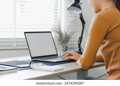 Businesswoman typing on a laptop with a blank screen at a desk in a well-lit home office - Powered by Shutterstock