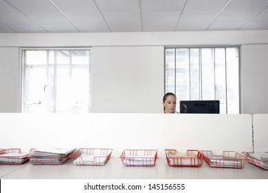 Businesswoman Using Computer In Office Cubicle Behind Trays With Documents