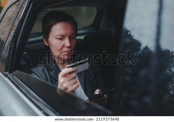 Businesswoman using credit card and mobile smart\
phone for e-banking from backseat of the car in rain, selective\
focus
