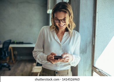 Businesswoman using her smart phone in office. Female entrepreneur looking at her mobile phone and smiling. Reading text messages. - Powered by Shutterstock